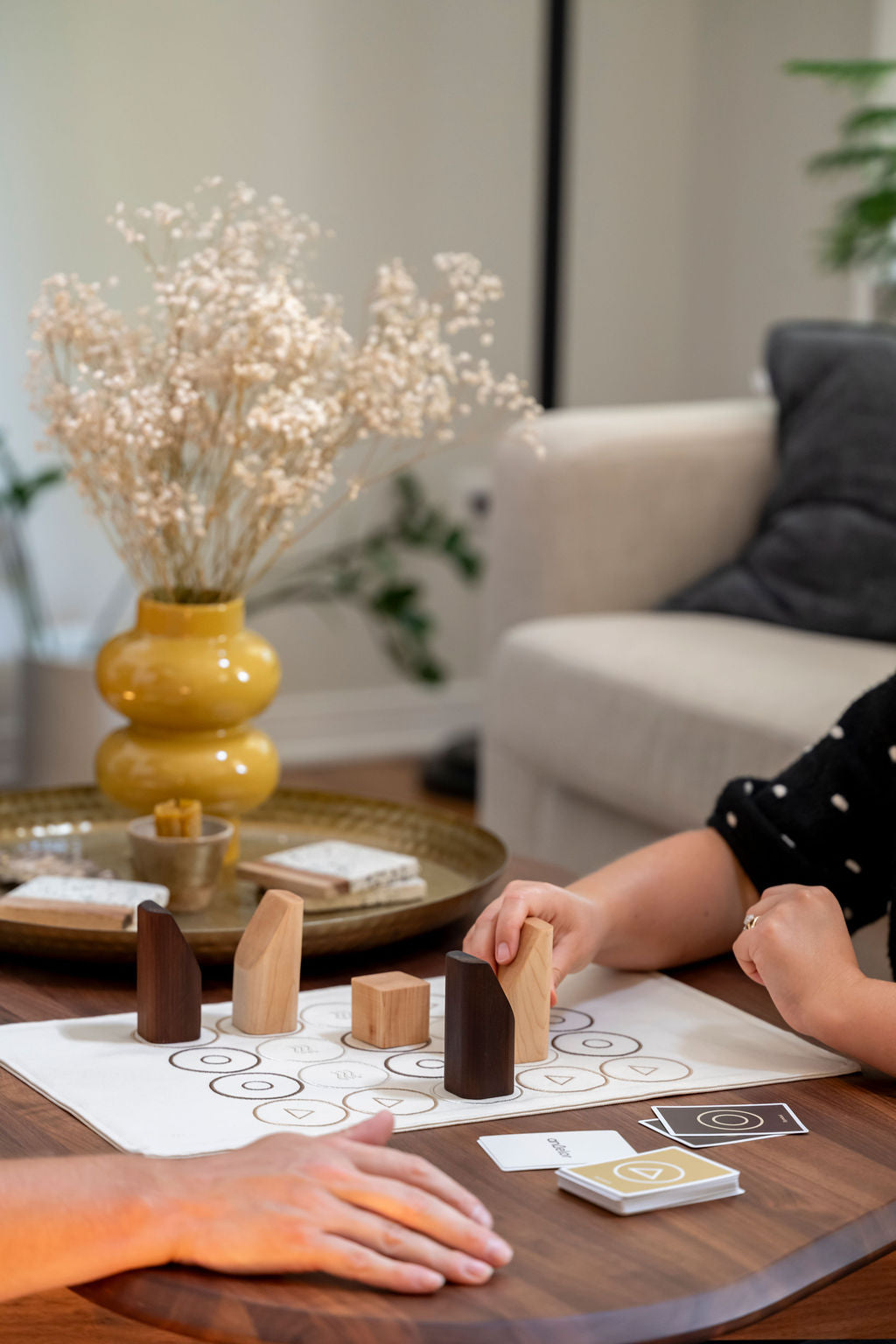 A close-up lifestyle shot of a wooden block game in progress on a wooden table. A person's hands are visible interacting with the game pieces, which include dark brown and light natural wood blocks arranged on a white game mat.